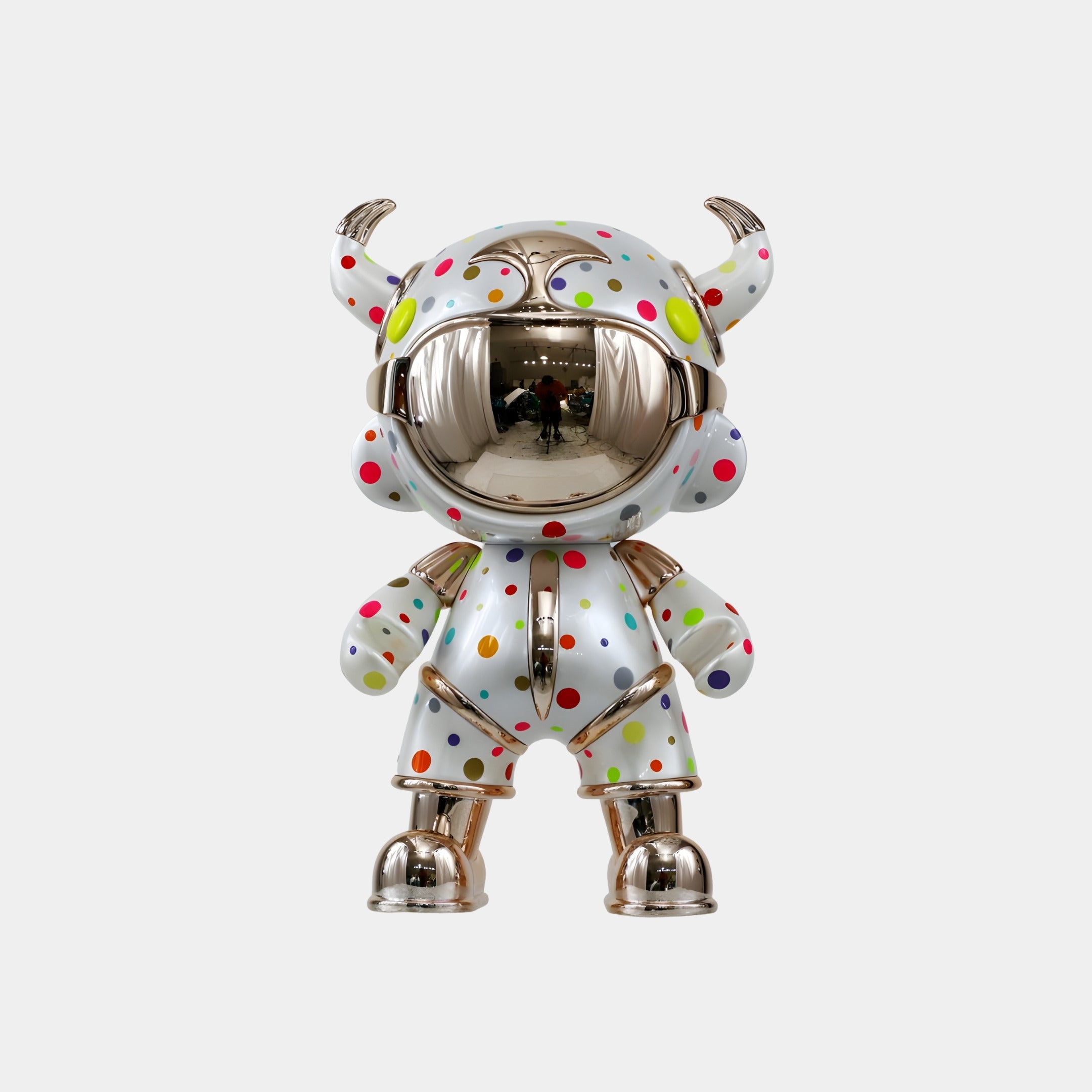 The White Confetti Suit Astro Devil Figure by Giant Sculptures stands 48cm tall, featuring a shiny metallic finish, round mirrored face, white suit with colorful polka dots, and horned helmet—a striking piece of contemporary art.