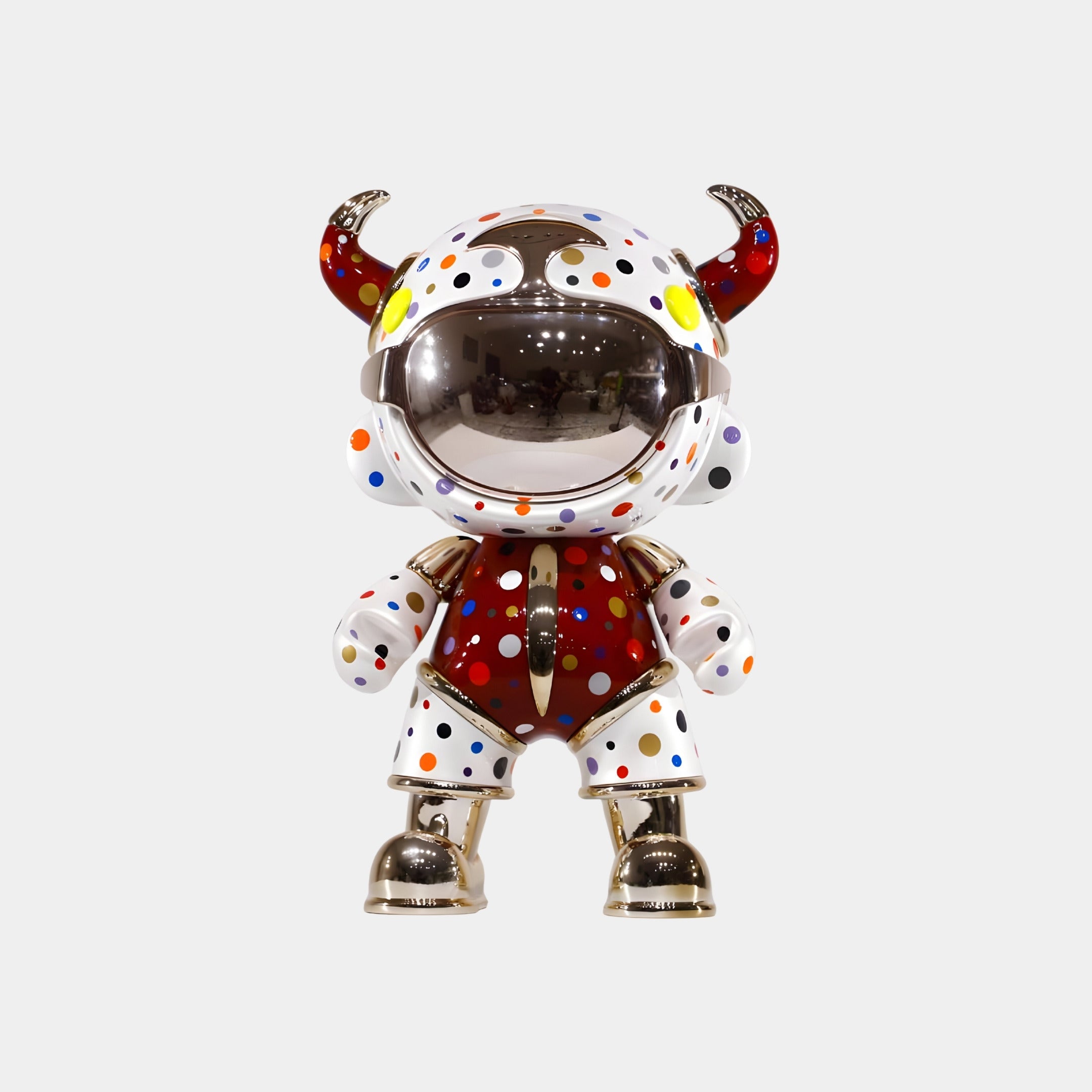 The Giant Sculptures Crimson Confetti Suit Astro Devil Figure (48cm) features a horned helmet, metallic gold accents, and mirrored visor. Its red body with white panels and colorful polka dots creates a playful pop-art look against a white background.