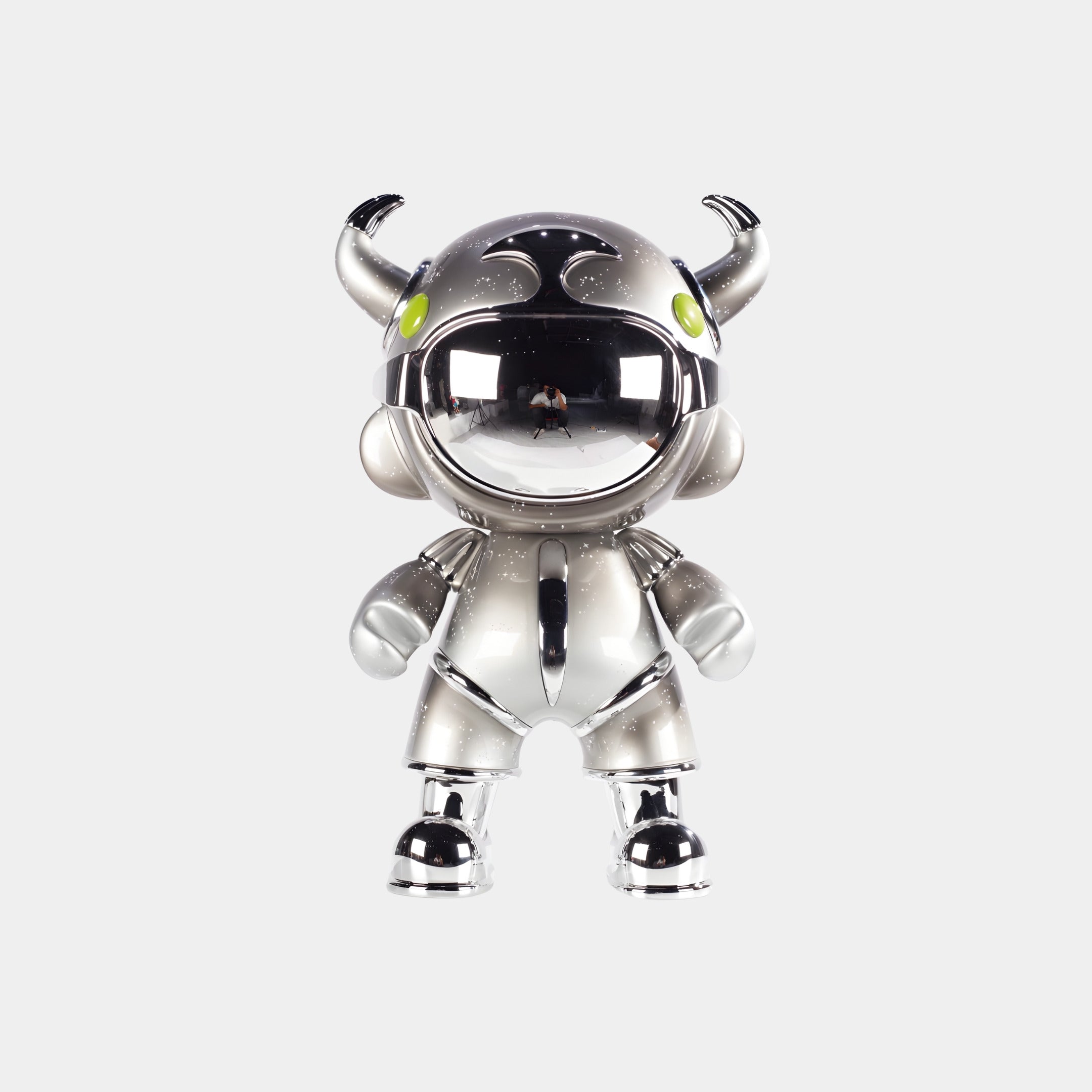The Platinum Suit Astro Devil Figure - 48cm by Giant Sculptures features a shiny, metallic finish, bull-like horns, green eyes, and round details. It stands upright with a mirrored visor against a plain white background.