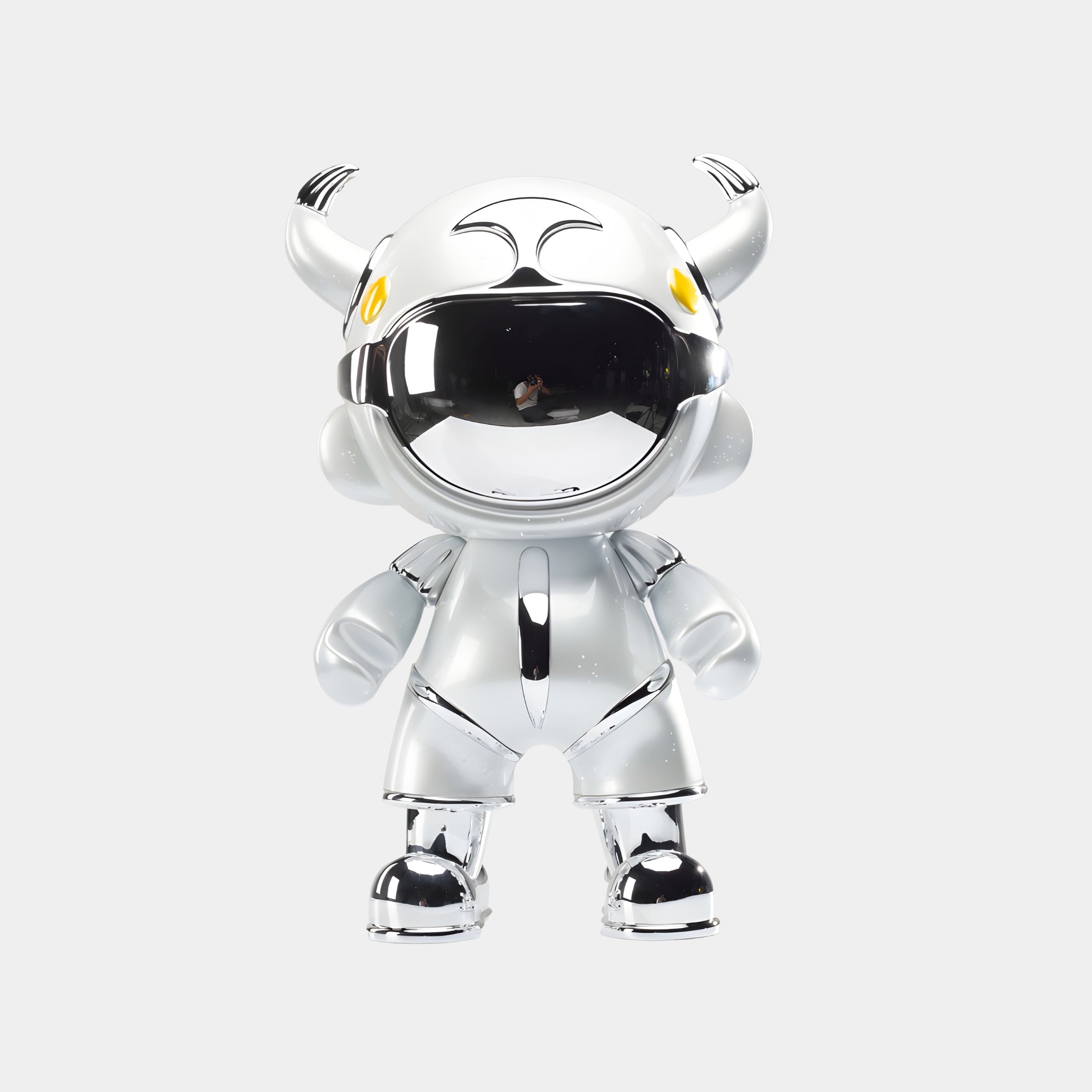 The Giant Sculptures Silver Suit Astro Devil Sculpture (80cm) features a shiny silver finish, mirrored chrome visor, large helmet with two small horns, yellow eyes, and rounded limbs, standing upright against a plain white background.