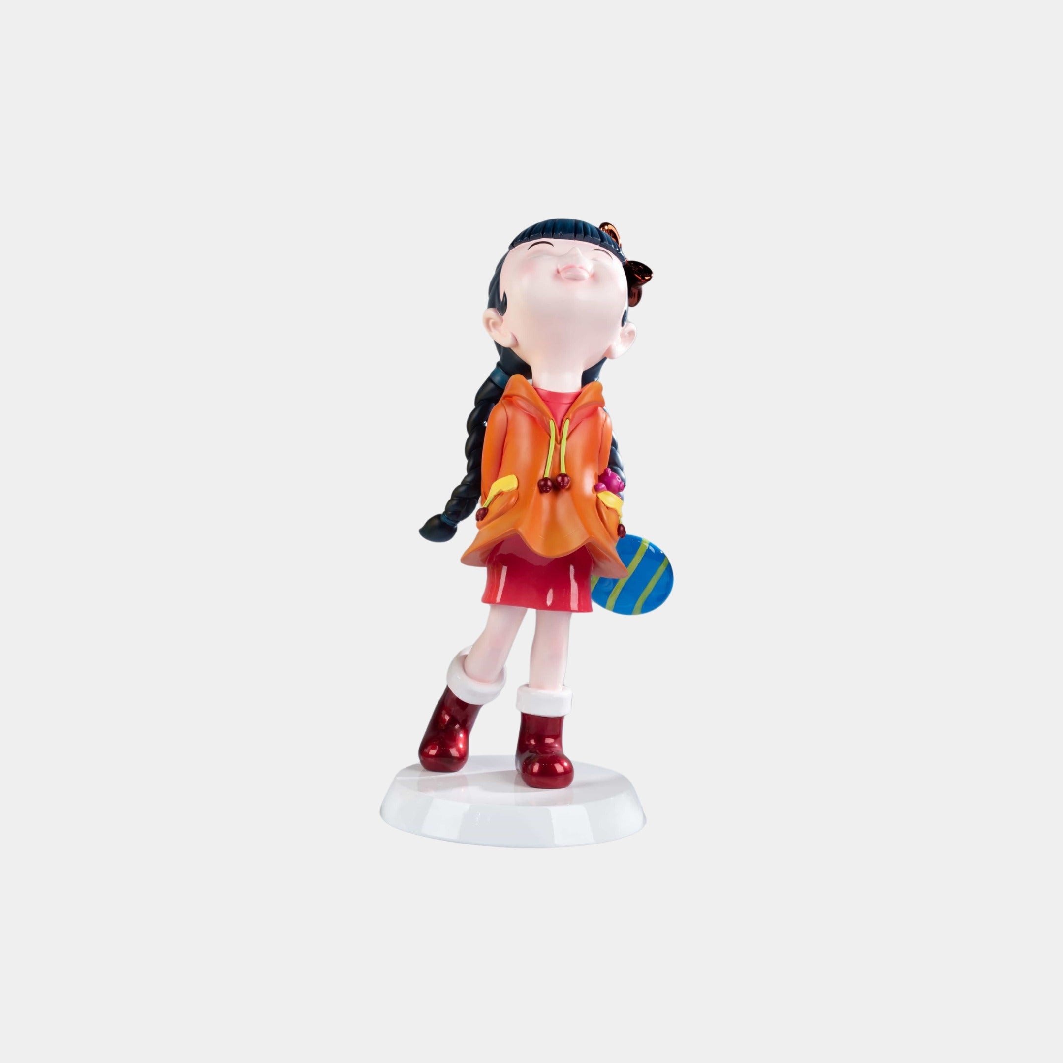 The Tangerine Orange Lulabelle Figure by Giant Sculptures is a 20cm eco-friendly decorative piece featuring a girl with black braided hair in an orange jacket and red skirt, standing on a white base while holding a striped ball behind her back.