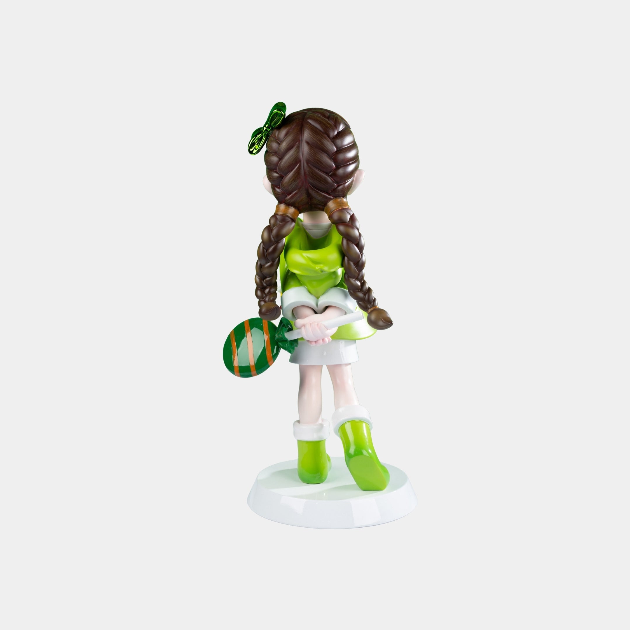 The Meadow Green Lulabelle Sculpture - 60cm by Giant Sculptures depicts a girl in green with braided brown hair and a striped paddle, crafted from eco-friendly polyurethane on a white base, facing away from the viewer.