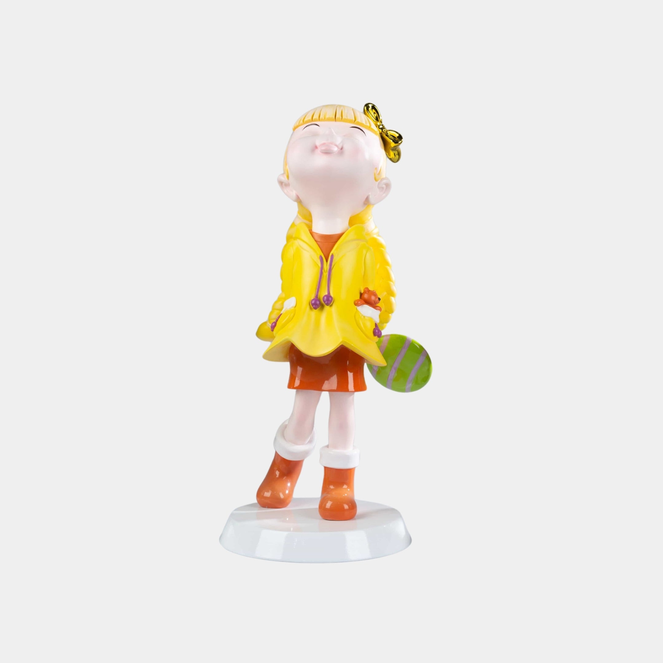 The Sunbeam Yellow Lulabelle Sculpture (60cm) by Giant Sculptures features a joyful girl in an orange skirt and boots, yellow jacket, and butterfly on her nose, holding a green-striped object—a charming décor piece on a white base.