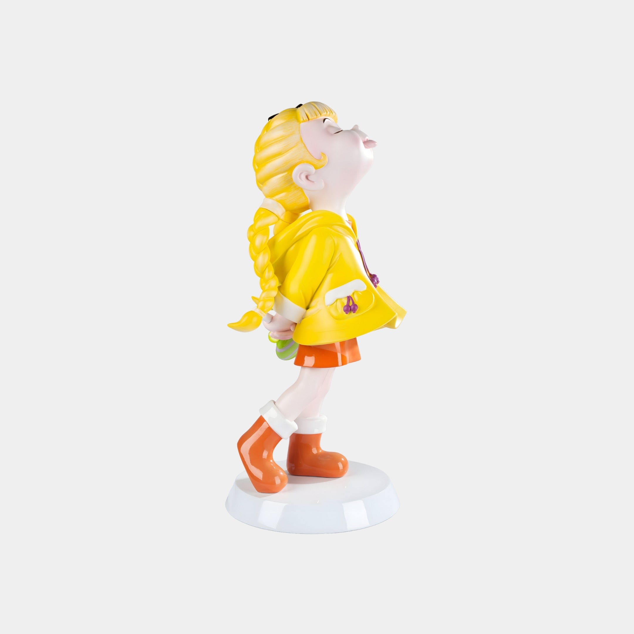The Sunbeam Yellow Lulabelle Sculpture - 60cm by Giant Sculptures features a joyful girl with blond braids in a yellow raincoat and orange boots, holding something behind her back on a white circular base.