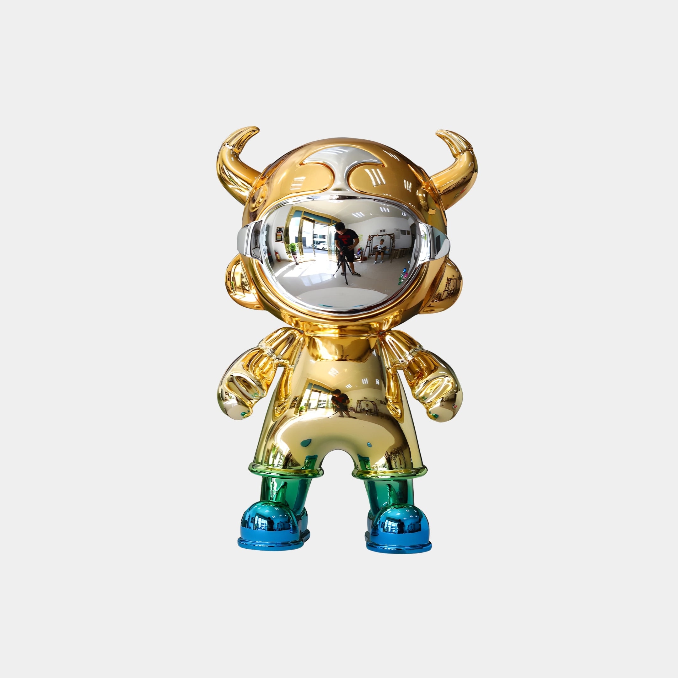 The Gold & Teal Electroplated Astro Devil Figure (35cm) by Giant Sculptures features a shiny gold helmet with distorted reflections, horns, green legs, and blue shoes, standing against a plain white background.