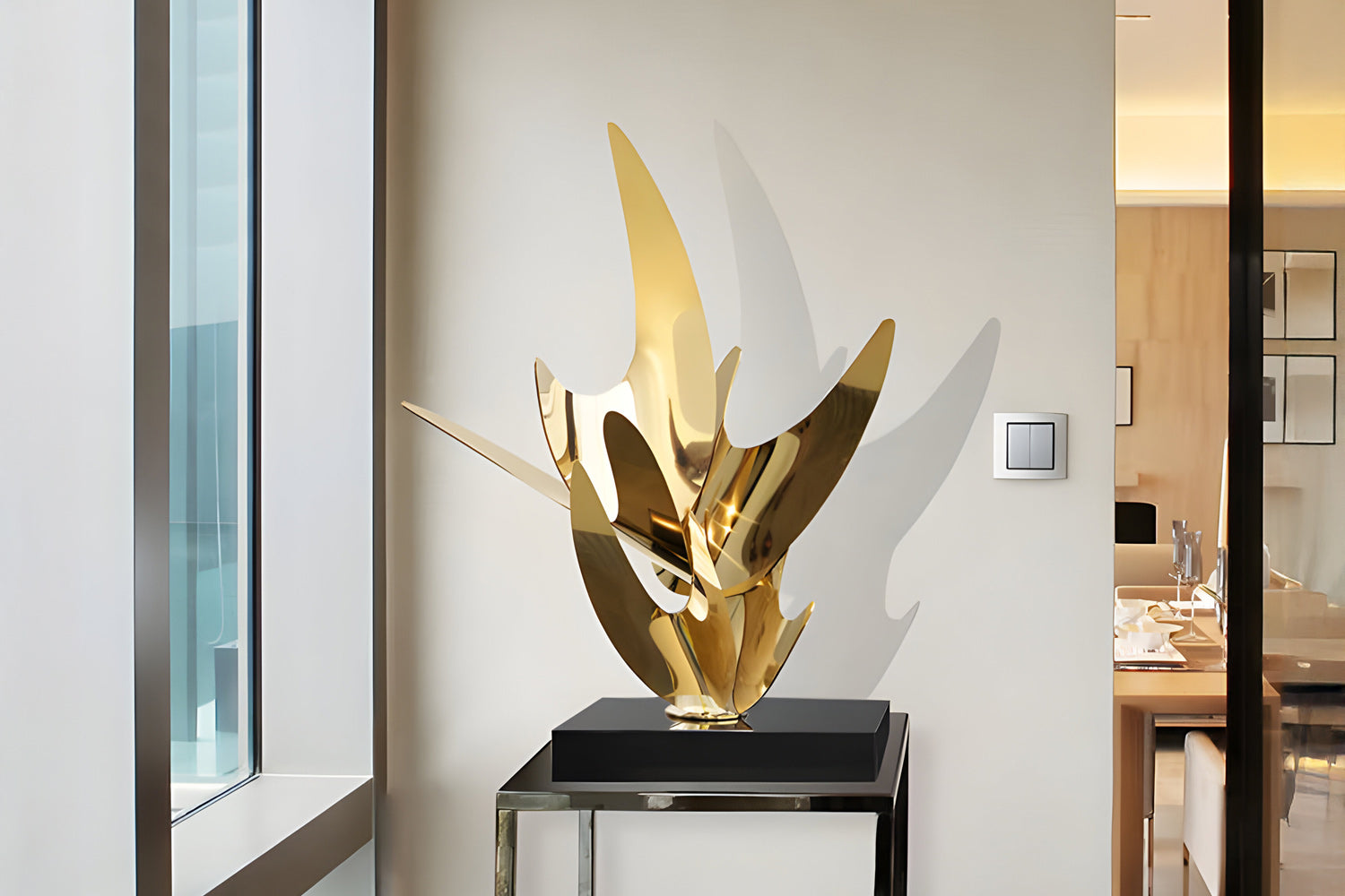 A modern, abstract gold sculpture on a black pedestal is placed in a bright room with large windows. The room has a minimalist design with light-colored walls and a glimpse of a dining area with simple decor in the background.