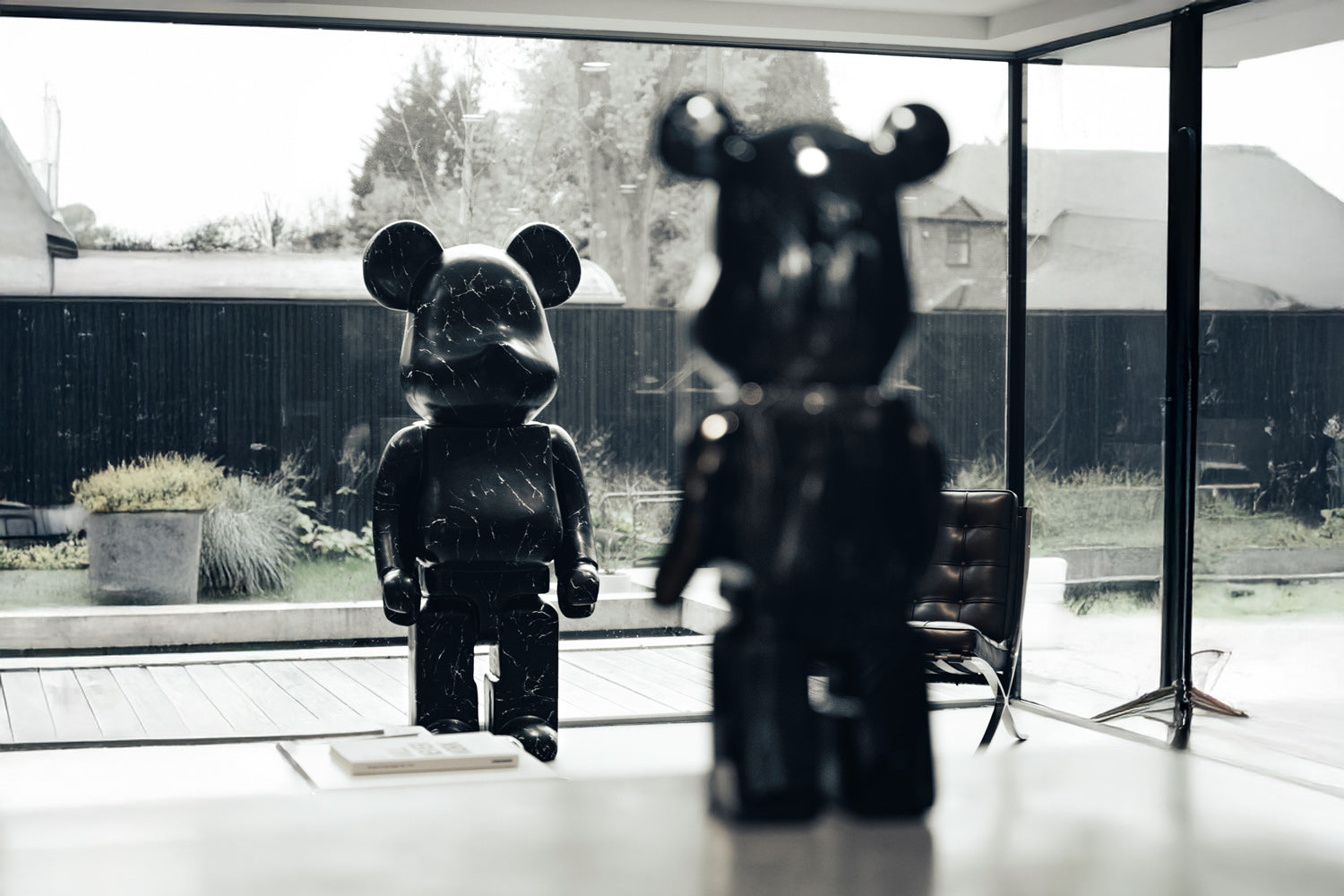 Two large, black bear sculptures with a glossy finish are positioned in a modern living space with floor-to-ceiling windows. One is in the foreground, while the other stands near the outdoor area, with greenery visible outside.