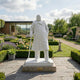 The Winston Churchill Marble Outdoor Sculpture - 195cm by Giant Sculptures depicts Churchill in a long coat with a cane, positioned in a landscaped garden with hedges, flowers, benches, and a sundial before a modern glass building.