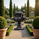Classical Bronze Cherub Urn Sculpture by Giant Sculptures on a stone plinth in a formal garden with cypress trees, clipped hedging, white roses, and gravel paths.