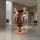 Giant Sculptures Classical Bronze Urn Vase with Figural Frieze, 100cm, on a red marble plinth in a neutral gallery space with archways.