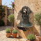 Classical Lion Head Bronze Fountain 70cm by Giant Sculptures mounted on a sandstone courtyard wall, water flowing from the lion's mouth into a scalloped basin beside potted lavender and olive trees.