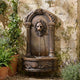 Classical Lion Head Fountain Bronze Sculpture by Giant Sculptures, 100cm wall-mounted piece with arched panel and basin, set against a weathered stone wall beside potted ferns and lavender.