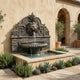 Giant Sculptures Classical Lion Head Wall Fountain in bronze with antique patina, mounted on a rendered courtyard wall beside olive trees, succulents, and stone arches.