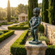 Classical Manneken Pis Boy Bronze Fountain by Giant Sculptures on a stone pedestal, water arcing into a formal garden with clipped hedges and cypress trees.