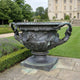 Classical Ornate Urn Bronze Sculpture by Giant Sculptures with bearded face medallions and scrolled handles, set on a terrace beside a manicured garden and manor.