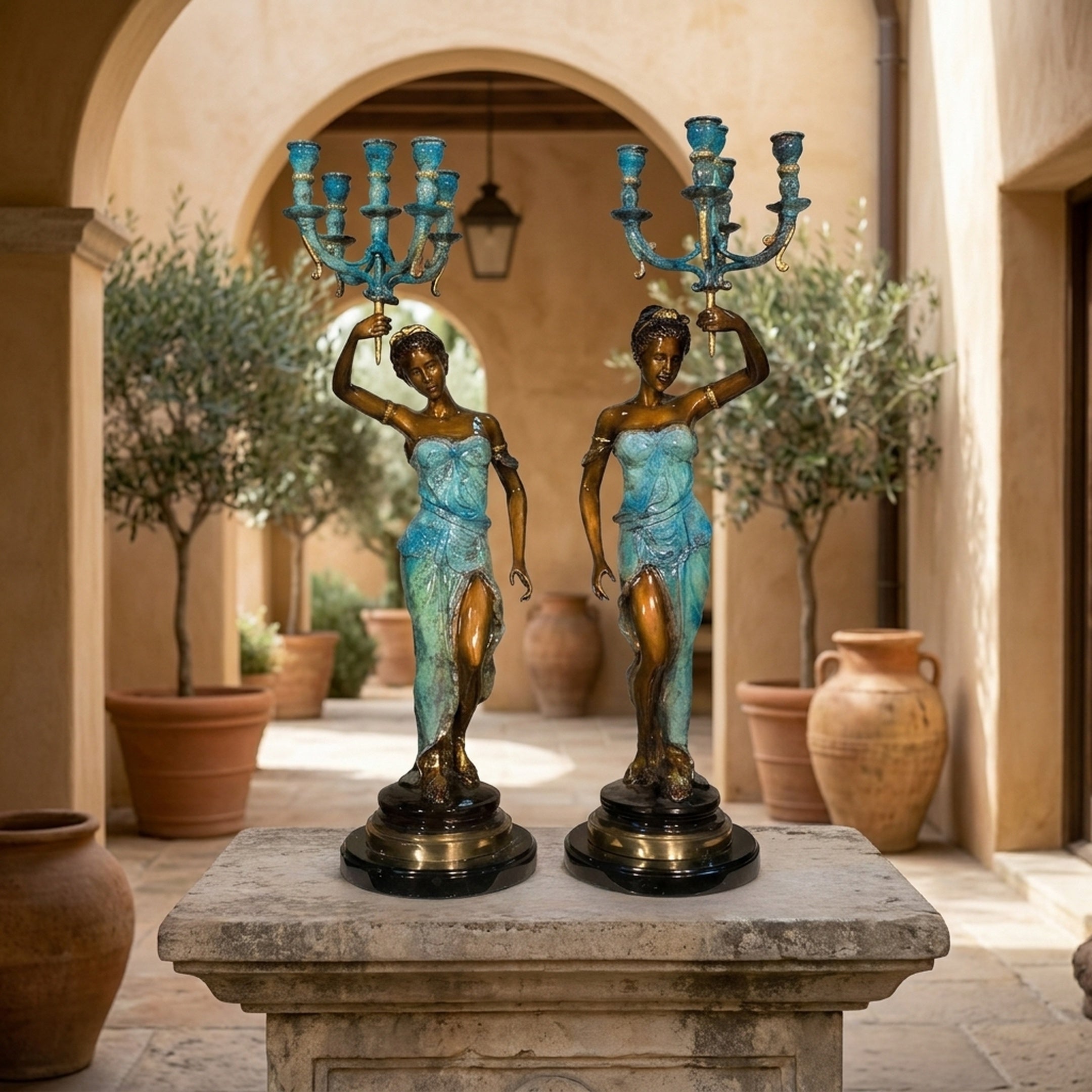 Classical Paired Goddess Candelabra Bronze Sculpture by Giant Sculptures, teal and gold patinated figures holding seven-arm candelabra, on a stone plinth in a Mediterranean courtyard.