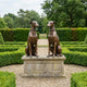 Classical Seated Greyhound Pair Bronze Sculpture by Giant Sculptures, two mirrored dogs with warm brown patina on a stone plinth in a formal hedged garden.