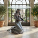 Classical Seated Mermaid Bronze Sculpture Table by Giant Sculptures, holding a conch beneath a round glass top, displayed in a sunlit conservatory with potted citrus trees.