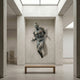 Contemporary Emerging Male Figure Bronze Wall Sculpture by Giant Sculptures, 90cm, mounted in a skylit gallery alcove above a wooden bench.