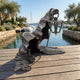 Contemporary Hooded Phantom Figure Bronze Sculpture by Giant Sculptures crawling on a timber dock, raised arm holding a lantern, with a marina behind.