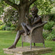 Giant Sculptures' 150cm Contemporary Reading Woman Bronze Sculpture, seated in a wicker armchair reading a book, displayed on a stone plinth beneath a garden tree.