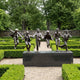Contemporary Running Children Bronze Sculpture by Giant Sculptures, six figures mid-stride on a dark plinth in a manicured boxwood garden with gravel paths.