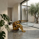 Contemporary Seated Nude Female Figure Bronze Sculpture by Giant Sculptures in golden-brown patina, supporting an oval glass tabletop in a minimalist room beside a courtyard olive tree.