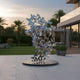 The Flutter Silver Butterfly Steel Sculpture - 200cm by Giant Sculptures features a silver butterfly design forming a human head on a circular base, set in a landscaped patio with green lawn, palm trees, and a modern building backdrop.
