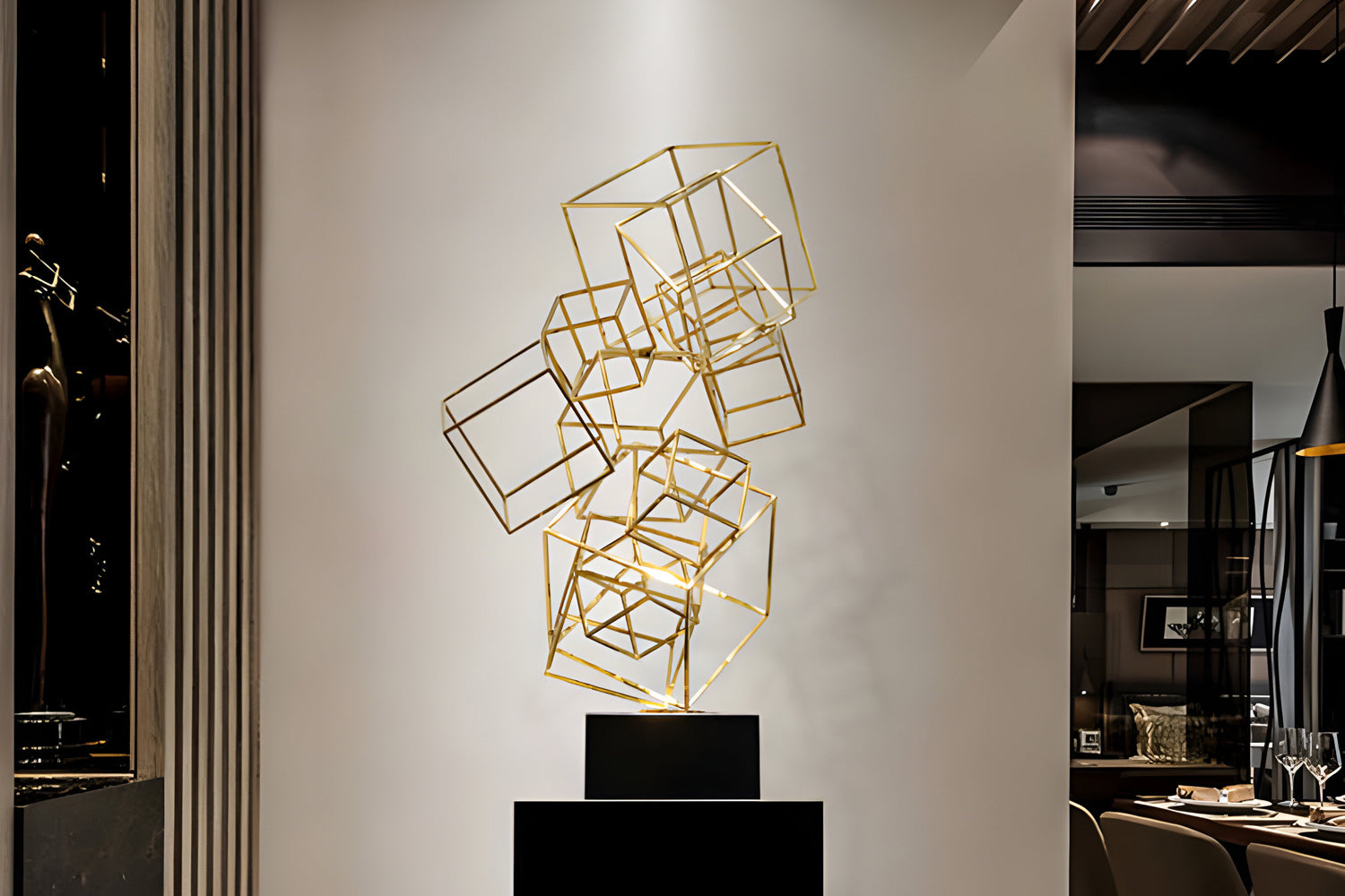 A sculpture of interconnected golden wireframe cubes is displayed on a black pedestal against a white wall. The modern art piece is set in an upscale interior with elegant decor and dining furniture visible in the background.