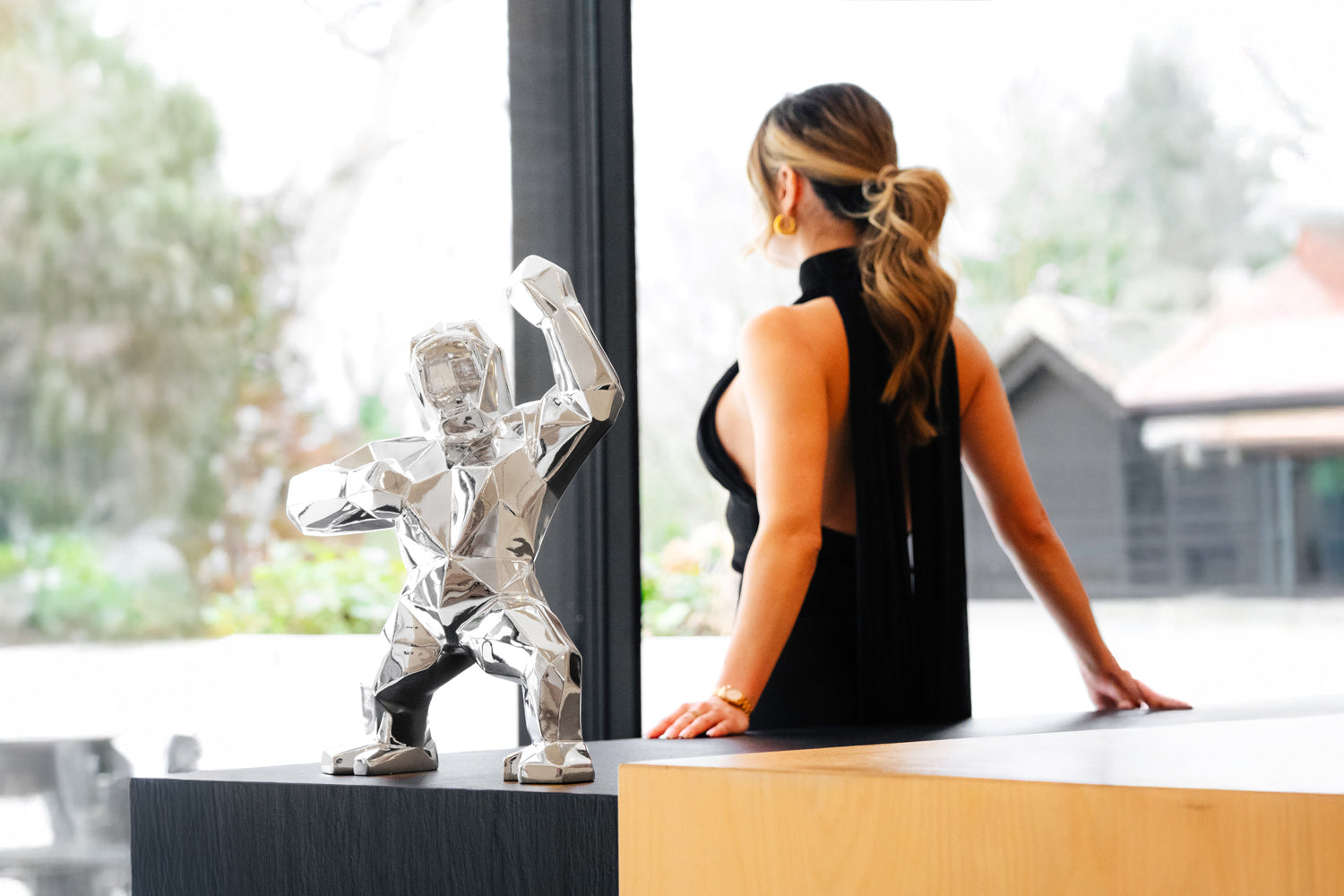 A woman with long hair in a ponytail stands inside, leaning against a countertop, wearing a black, sleeveless dress. In the foreground, a small, silver, geometric gorilla sculpture is displayed on the countertop.