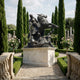 Giant Sculptures' 260cm Classical Armoured Knight and Horse Slaying Dragon bronze on a pale stone plinth, framed by cypress trees and rose bushes in a formal garden.