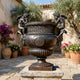 Large Classical Cherub Urn Bronze Sculpture by Giant Sculptures on a stone plinth in a Mediterranean courtyard with olive trees and bougainvillea.