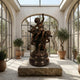 Large Classical Cherubs and Central Figure Bronze Sculpture by Giant Sculptures displayed on a stone plinth in a sunlit arched conservatory with olive trees.