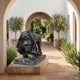 Large Classical Crouching Lion Bronze Sculpture by Giant Sculptures with dark patina and verdigris, displayed on a stone plinth in an arched Mediterranean courtyard.