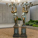 Pair of Giant Sculptures Large Classical Draped Goddess Candelabra Bronze Sculptures in verdigris and gold finish, displayed on ornate pedestals in a concrete courtyard.