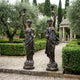 Large Classical Goddess Pair Bronze Sculpture by Giant Sculptures, 180cm draped female figures raising candelabra torchères, displayed on a formal garden path.