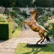 Giant Sculptures Large Classical Rearing Horse Bronze Sculpture 180cm with warm golden-amber patina on a dark rocky base, displayed on a garden path beside flowerbeds.