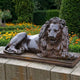 Giant Sculptures Large Classical Recumbent Lion Bronze Sculpture in dark patina, resting on a plinth atop a stone wall beside marigolds and hedges.