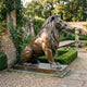 Large Classical Seated Lion Bronze Sculpture by Giant Sculptures shown in profile with warm reddish-brown patina on a low plinth beside a walled garden pond.