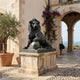 Large Classical Seated Lion Bronze Sculpture by Giant Sculptures in dark antique patina, left paw resting on a sphere, on a stone plinth in a sunlit Mediterranean courtyard.