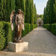 Large Classical Standing Angel Bronze Sculpture by Giant Sculptures on a stone plinth along a gravel path lined with tall cypress hedges leading to a pavilion.