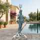 Large Classical Standing Poseidon Bronze Sculpture by Giant Sculptures with verdigris patina, holding a trident on a stone base beside a Mediterranean pool.