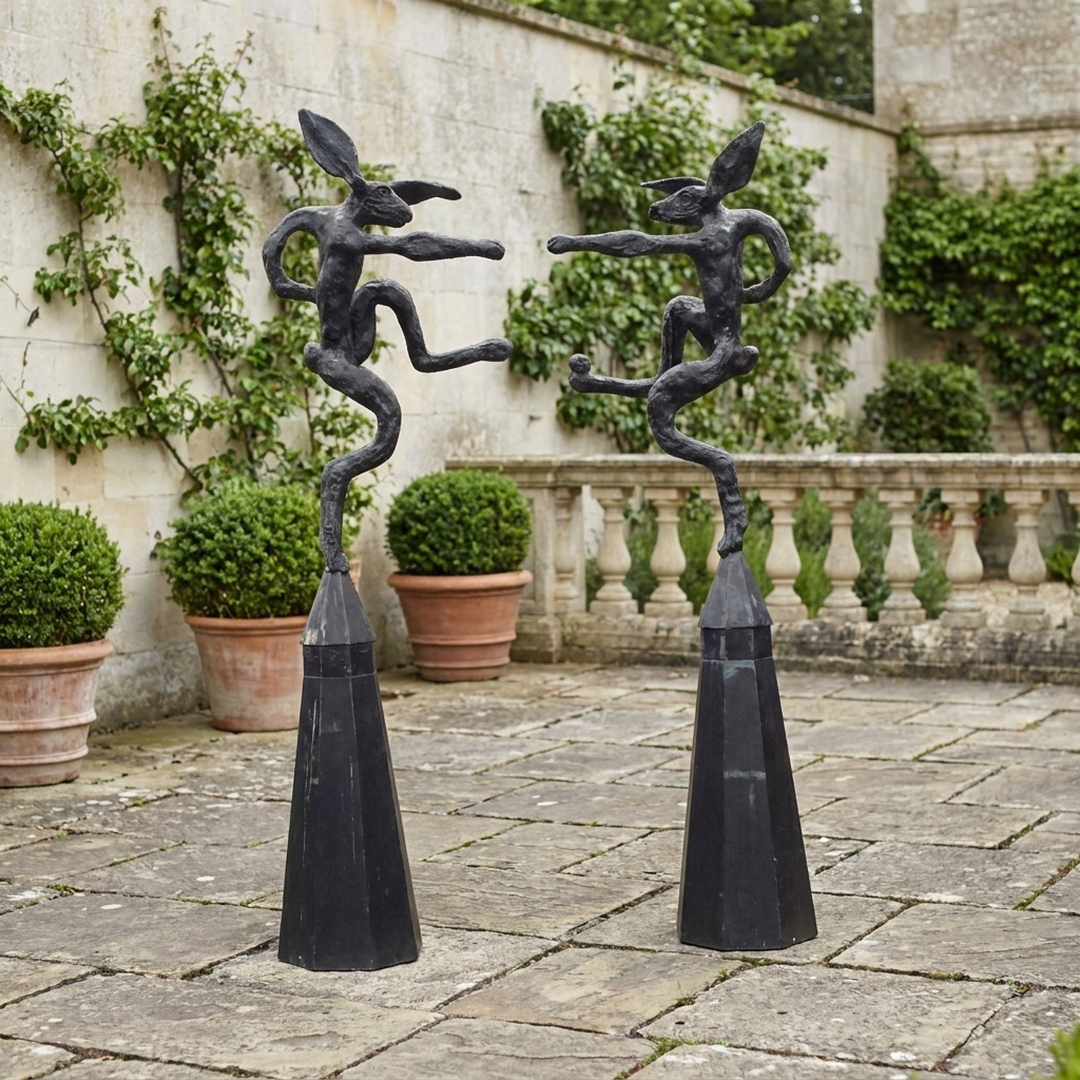 Giant Sculptures Large Contemporary Fighting Hares Bronze Sculpture 180cm, two dark patinated hares squaring off on tapered obelisk plinths in a garden terrace.