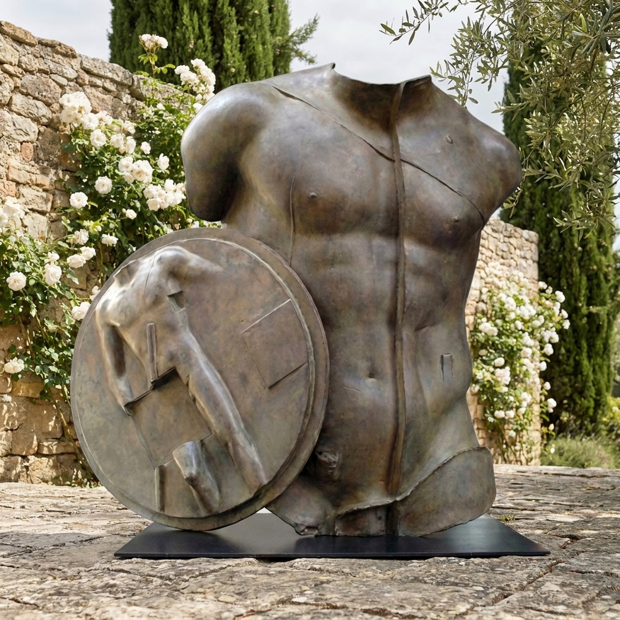 Large Contemporary Fragmentary Warrior Torso Bronze Sculpture by Giant Sculptures, displayed outdoors on stone paving beside white roses and cypress trees.