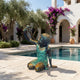 Large Contemporary Girl with Bird Bronze Sculpture by Giant Sculptures, seated girl lifting a bird skyward, verdigris and golden patina, beside a poolside courtyard.