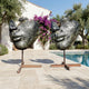 Large Modern Abstract Face Fragment Bronze Sculpture by Giant Sculptures, two 180cm cracked olive-brown face fragments on steel poles beside a poolside patio.
