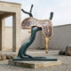 Large Modern Melting Clock Bronze Sculpture by Giant Sculptures, 220cm, with golden clock face draped over a green-black twisted trunk in a gravel courtyard.