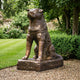Large Traditional Akita Dog Bronze Sculpture by Giant Sculptures, seated with head raised on a textured plinth, set on gravel beside a lawn and garden hedging.