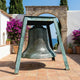 Large Traditional Bronze Bell Sculpture by Giant Sculptures, 90cm, hanging in a verdigris A-frame stand on a tiled Mediterranean terrace with bougainvillea and sea view.