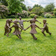 Large Traditional Dancing Children Bronze Sculpture by Giant Sculptures, six children holding hands in a circle on a garden lawn with flowering shrubs behind.